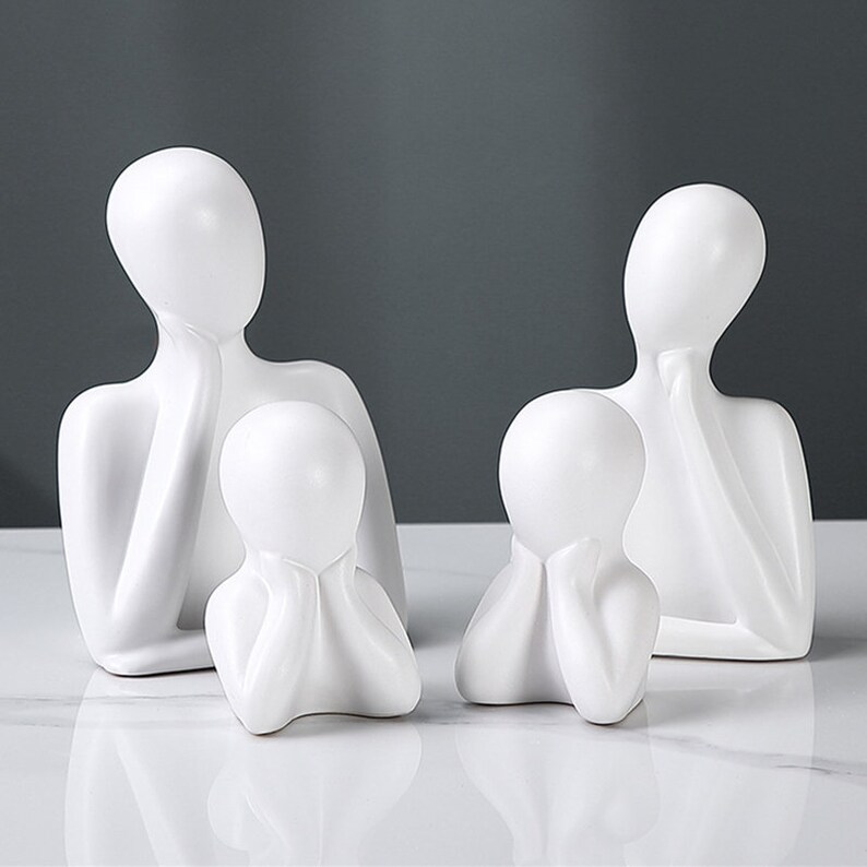May include: Four white ceramic sculptures depicting abstract human figures. Two larger figures stand behind two smaller figures, all with a minimalist design. The sculptures have smooth surfaces and are set against a neutral background.