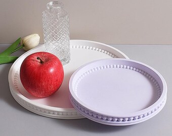 May include: A still-life composition featuring a red apple, a clear glass vase, and two decorative trays. One tray is white, and the other is lavender. The trays have a beaded edge design. A white tulip is also visible.
