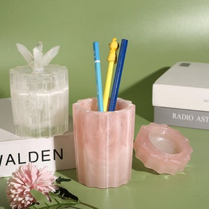 May include: Three decorative containers made of translucent pink and white resin with a marbled effect. The containers have a starburst design on the lid and a fluted design on the sides. The containers are filled with pencils and a small figurine.