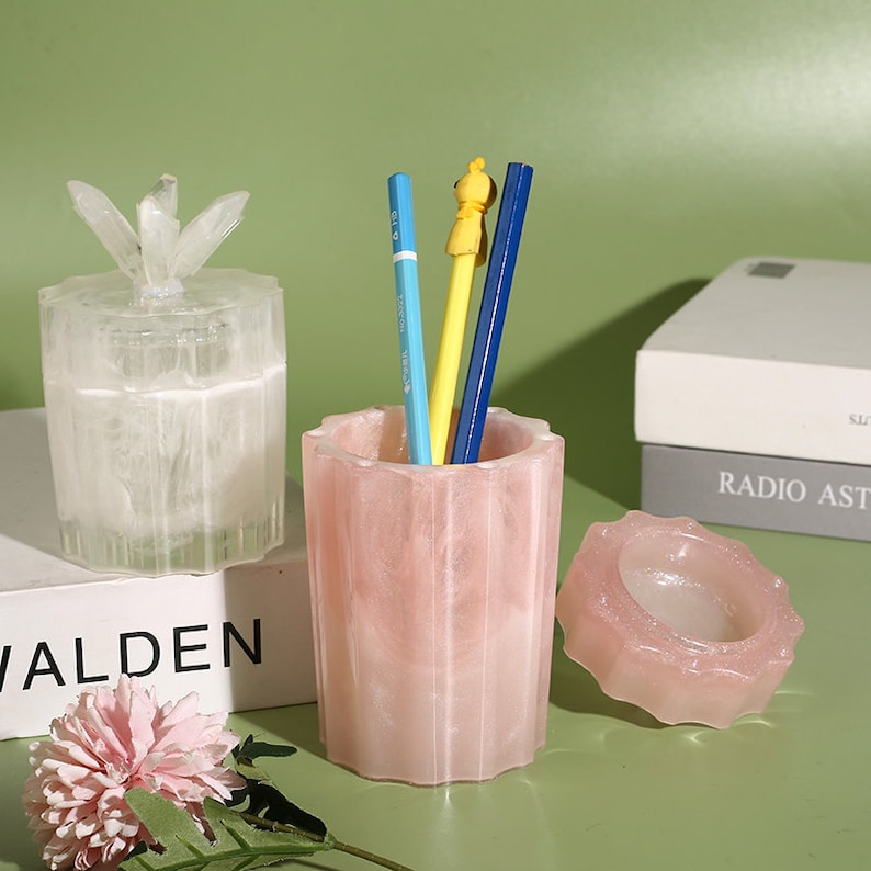 May include: Three decorative containers made of translucent pink and white resin with a marbled effect. The containers have a starburst design on the lid and a fluted design on the sides. The containers are filled with pencils and a small figurine.