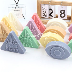 Puede incluir: Surtido de velas coloridas en varias formas, incluyendo triángulos y óvalos. Cada vela presenta un diseño de ojo estilizado con líneas radiantes. Las velas están dispuestas sobre una superficie blanca.