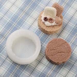 May include: A white silicone mold for making round cakes, next to a finished cake decorated with white chocolate, cereal, and a cookie.