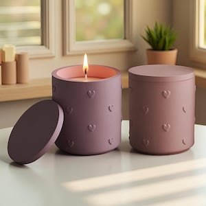 Puede incluir: Dos velas cilíndricas con diseños en relieve en forma de corazón. Una vela está encendida, con interior rosa y mecha encendida. La otra vela tiene una tapa a juego. Las velas son de color púrpura apagado y están sobre una superficie blanca.