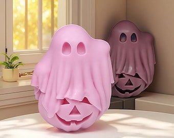Puede incluir: Dos decoraciones temáticas de Halloween. El primero es un fantasma rosa claro con una cara de calabaza tallada. El segundo es un fantasma morado más oscuro con un diseño similar. Ambos están sobre una superficie blanca cerca de una ventana.