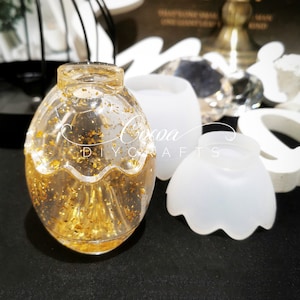 Puede incluir: Un recipiente de vidrio transparente con forma de huevo lleno de purpurina dorada. El recipiente está sobre una superficie negra junto a un molde de silicona blanco en forma de huevo.