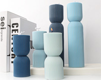 May include: A collection of pillar candles in various shades of blue and green. The candles have a unique, two-part design with vertical ribbed detailing. One candle is lit, showing a small flame.