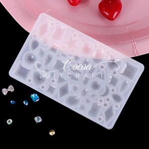 May include: A clear silicone mold with 24 different shapes of gemstones, hearts, and other geometric shapes. The mold is on a pink plate. The text "Cocoa DIY Crafts" is visible on the mold.