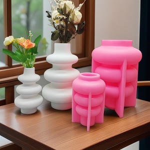 May include: A collection of decorative vases in various sizes and colors. Two white, stacked-sphere vases hold flowers. Two pink vases, one with a unique structural design, sit on a wooden table. The vases are a modern home decor accent.