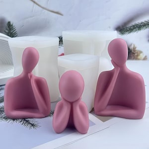 May include: Three pink human figure sculptures in various poses, with hands supporting the head or chin. Behind the sculptures are white, rectangular molds. The sculptures are on a light-colored surface.