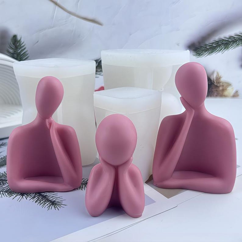 May include: Three pink human figure sculptures in various poses, with hands supporting the head or chin. Behind the sculptures are white, rectangular molds. The sculptures are on a light-colored surface.