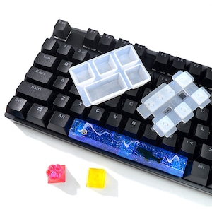 May include: A black keyboard with a blue and white resin keycap, a clear silicone mold for making keycaps, and two small silicone molds for making keycaps. The molds are on a white surface.