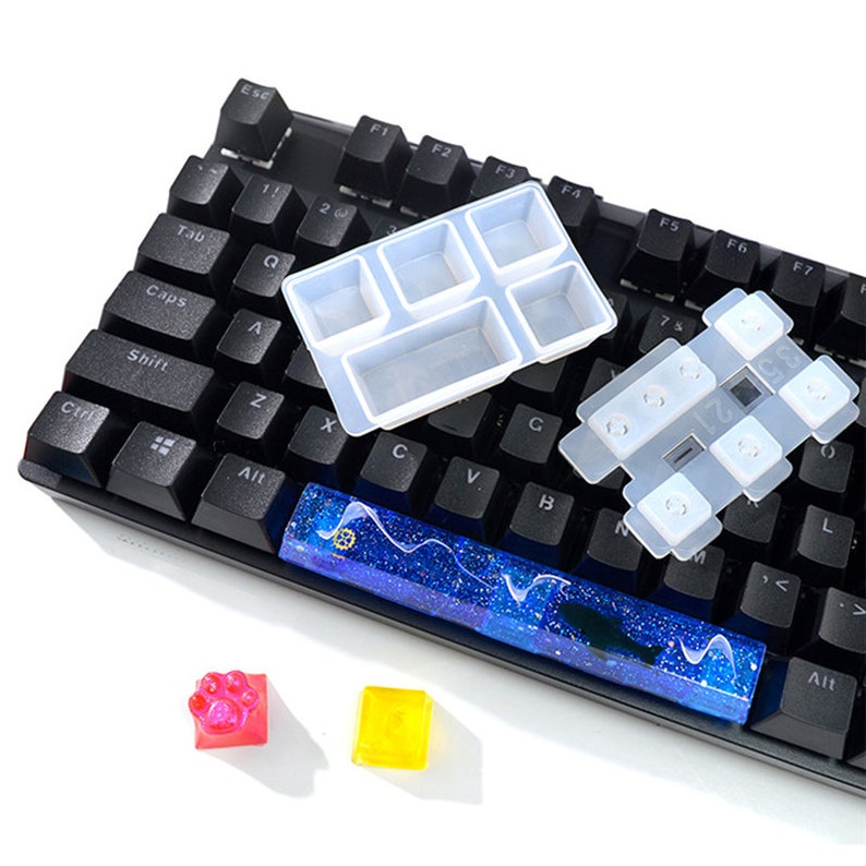 May include: A black keyboard with a blue and white resin keycap, a clear silicone mold for making keycaps, and two small silicone molds for making keycaps. The molds are on a white surface.