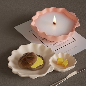 Könnte beinhalten: Eine rosa Kerze mit gerüschtem Rand in einer Kuchenform mit einem brennenden Docht. Eine weiße, wellenförmige Schale enthält ein Brötchen mit Butter und zwei gelbe Butterwürfel. Ein silbernes Messer liegt daneben.