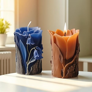 May include: Two decorative candles. One is blue with a floral design, the other is orange and lit. Both candles have a wood-like texture around the base. The blue candle features a flower and bell-shaped blossoms.