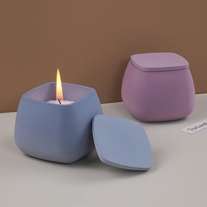 May include: Two square-shaped candles in pastel colors. One is lit with a flame, the other has a lid. The candle containers are blue and purple, with a matching lid. The candles are on a white surface with a brown background.