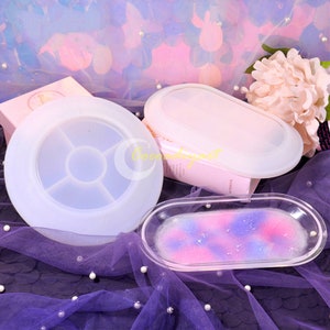 Op de afbeelding: Twee transparante siliconen mallen voor hars ambachten. Eén mal is rond met zes compartimenten en de andere is ovaal. De ovale mal wordt getoond met een afgewerkt hars dienblad met roze, blauwe en paarse swirls.