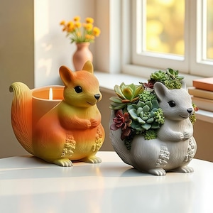 Puede incluir: Dos objetos decorativos con forma de ardilla. Uno es un portavelas en tonos naranja y amarillo. El otro es una maceta llena de suculentas. Ambos están sobre una superficie blanca, cerca de una ventana.