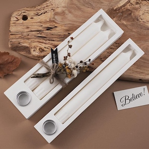 Peut inclure: Deux bougies coniques blanches dans une boîte blanche avec une base argentée. La boîte est décorée de fleurs séchées et attachée avec de la ficelle. Une petite carte avec le mot "Believe!" est également présente.