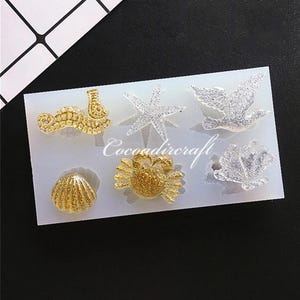 Peut inclure: Un moule en silicone avec six formes différentes sur le thème de la mer : une étoile de mer, un hippocampe, un crabe, une coquille, une coquille à bord festonné et une colombe. Le moule est blanc et les formes sont remplies de paillettes.
