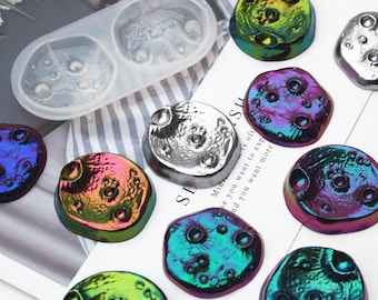 May include: A silicone mold with two cavities shaped like a moon with craters. There are multiple iridescent resin castings of the moon in various colors, including green, blue, purple, and silver.