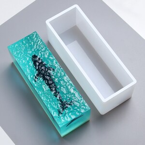 Puede incluir: Una pieza de arte rectangular de resina con un diseño de pez negro encerrado en un bloque de resina turquesa texturizada. Al lado hay un molde rectangular blanco vacío. La pieza de arte mide aproximadamente 15 cm de largo.