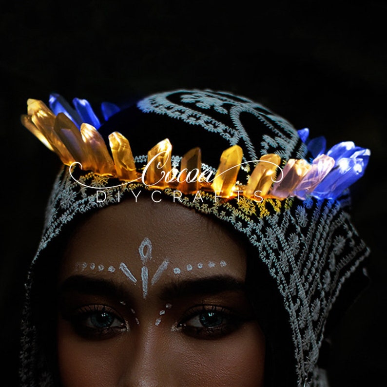 May include: A person wearing a black and white patterned head covering with a crown of glowing blue and yellow crystal-shaped lights. The text "Cocoa DIY Crafts" is visible on the head covering.