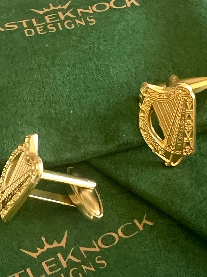 May include: A pair of gold cufflinks featuring a detailed design of a harp, the national symbol of Ireland. The cufflinks are displayed on a green velvet background with the text "Castle Knock Designs" printed on the surface.