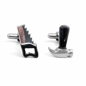 Könnte beinhalten: Ein Paar silberne Manschettenknöpfe, einer in Form einer Säge und der andere in Form eines Hammers. Die Säge hat einen schwarzen Griff und eine silberne Klinge. Der Hammer hat einen schwarzen Griff und einen silbernen Kopf. Auf weißem Hintergrund.