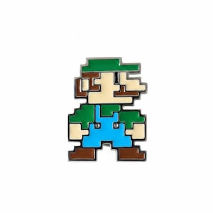 Könnte beinhalten: Eine verpixelte Emaille-Anstecknadel, die eine Figur mit grüner Mütze, braunen Haaren, cremefarbenem Gesicht, grünem Oberteil, hellblauer Latzhose und braunen Schuhen zeigt. Das Design ist mit silbernem Metall umrandet und wirkt wie ein Retro-Spiel.