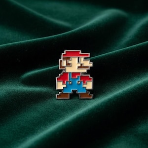 Könnte beinhalten: Eine Metall-Emaille-Anstecknadel mit einem verpixelten Charakterdesign. Die Figur trägt eine rote Mütze, ein rotes Oberteil, eine blaue Latzhose und braune Schuhe, mit beigem Gesicht und Händen. Das Design ist im blockartigen 8-Bit-Stil gehalten.