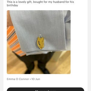 May include: Gold-coloured cufflinks featuring a detailed harp design. The cufflinks are shown on a light blue shirt with a striped orange and white tie.
