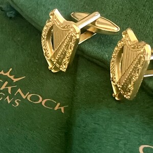 May include: Gold cufflinks featuring a detailed design of the Irish harp, a traditional symbol of Ireland. The cufflinks are displayed on a green velvet background.