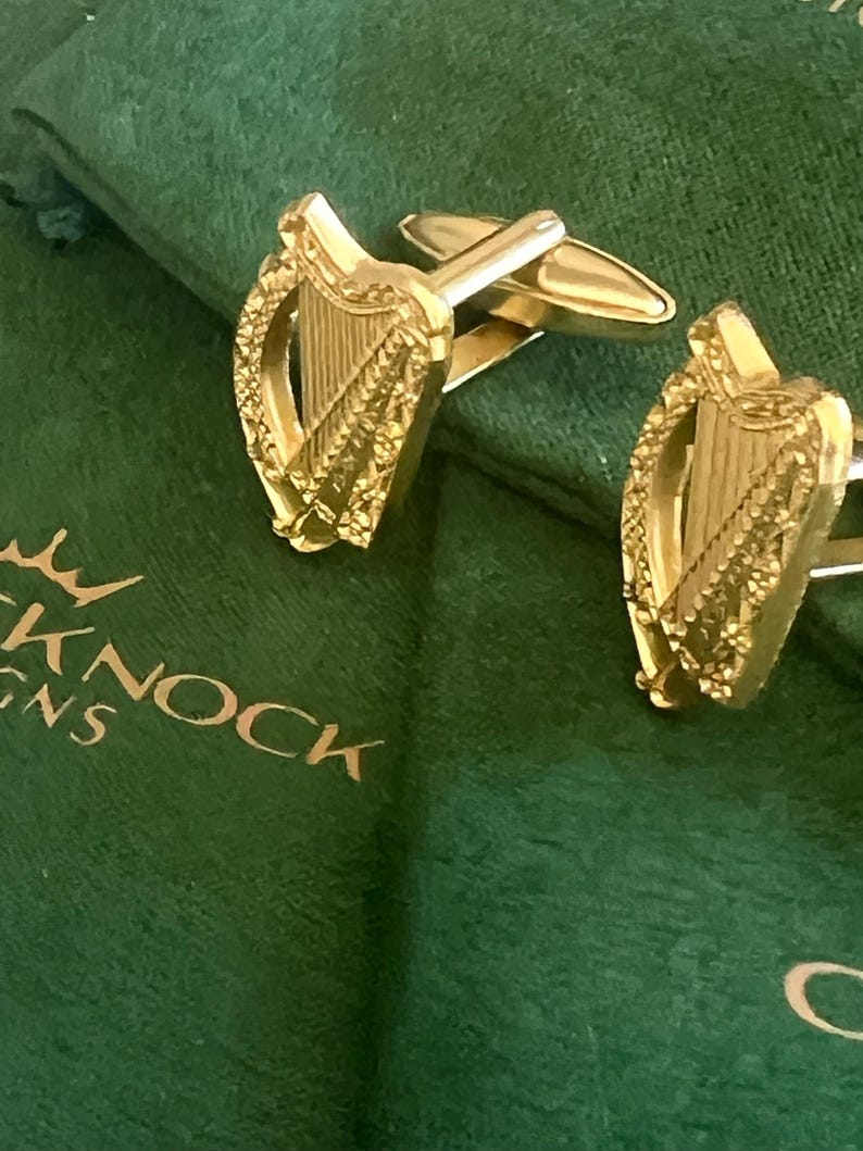 May include: Gold cufflinks featuring a detailed design of the Irish harp, a traditional symbol of Ireland. The cufflinks are displayed on a green velvet background.