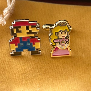 May include: A pair of gold cufflinks featuring pixelated characters from the Super Mario Bros. video game. The cufflinks are on a yellow velvet background. The left cufflink features Mario, a red-clad character with a mustache, and the right cufflink features Princess Peach, a pink-clad character with a crown.