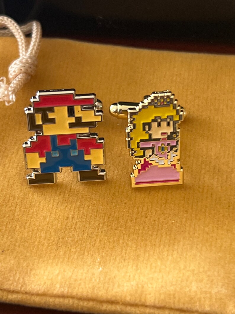 May include: A pair of gold cufflinks featuring pixelated characters from the Super Mario Bros. video game. The cufflinks are on a yellow velvet background. The left cufflink features Mario, a red-clad character with a mustache, and the right cufflink features Princess Peach, a pink-clad character with a crown.