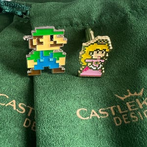 Könnte beinhalten: Ein Paar goldene Manschettenknöpfe mit pixelartigen Figuren aus dem Videospiel Super Mario Bros. Die Manschettenknöpfe befinden sich auf einem grünen Samt-Hintergrund mit dem Text "CASTLE ROCK DESIGNS".