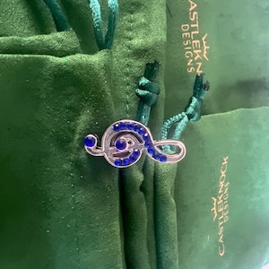 Könnte beinhalten: Ein silberner Violinschlüssel-Anhänger mit blauen Strasssteinen, der auf einem grünen Samtbeutel mit dem aufgedruckten Text "CASTLEKNOCK DESIGNS" liegt.