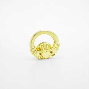 Könnte beinhalten: Goldfarbene Claddagh-Anstecknadel mit einem Herz, einer Krone und Händen. Das Design ist kreisförmig mit einem Stift auf der Rückseite. Das Herz wird von einer Krone gekrönt und von zwei Händen gehalten. Der Hintergrund ist weiß.