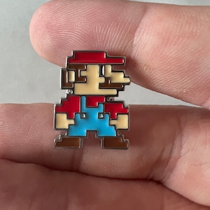 Peut inclure: Une épingle en émail pixelisée du personnage emblématique de jeu vidéo, Super Mario. L'épingle représente Mario dans sa salopette rouge et bleue classique, avec une moustache brune et un chapeau rouge.