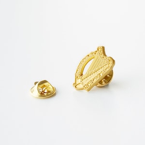 Könnte beinhalten: Goldfarbener Metallstift mit einem detaillierten Design einer Harfe, einem traditionellen irischen Symbol. Der Stift hat einen Sicherheitsverschluss auf der Rückseite.