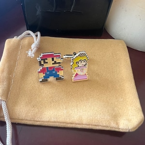 May include: A pair of gold-toned cufflinks featuring pixelated images of Mario and Princess Peach from the Super Mario Bros. video game. The cufflinks are displayed on a tan velvet pouch.