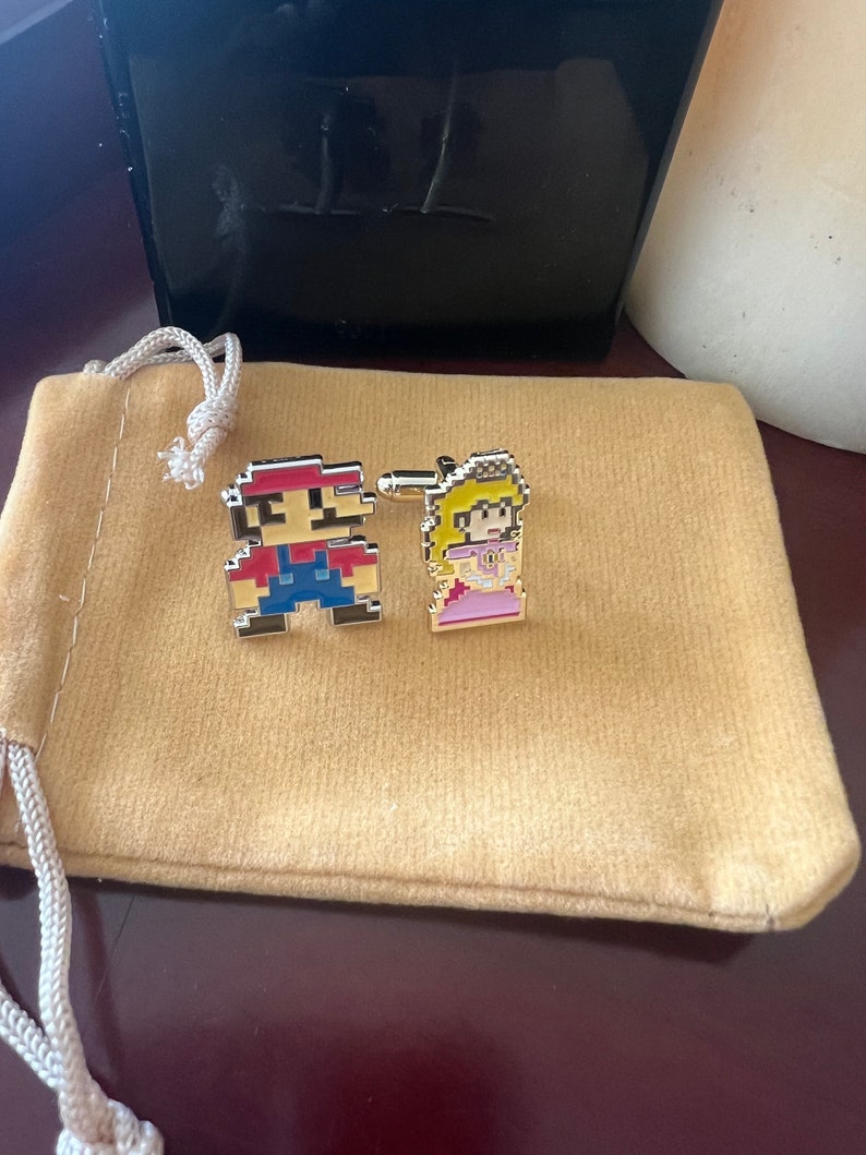 May include: A pair of gold-toned cufflinks featuring pixelated images of Mario and Princess Peach from the Super Mario Bros. video game. The cufflinks are displayed on a tan velvet pouch.