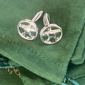 May include: A pair of silver cufflinks with a steering wheel design. The cufflinks are on a green velvet background.