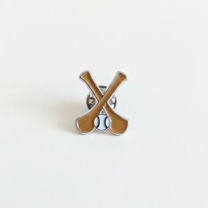 Könnte beinhalten: Eine Metall-Emaille-Anstecknadel mit zwei gekreuzten beigefarbenen Baseballschlägern und einem weißen Baseball in der Mitte am unteren Rand. Das Design hat eine silberfarbene Metallumrandung und einen sichtbaren Verschluss.