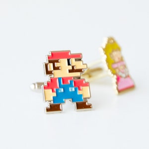 May include: A pair of cufflinks featuring a pixelated image of Mario from the Super Mario Bros. video game. The cufflinks are silver with red, blue, yellow, and white enamel.