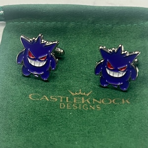 May include: A pair of cufflinks featuring a purple cartoon character with a wide grin and red eyes. The cufflinks are on a green drawstring bag with the text "CASTLEKNOCK DESIGNS".