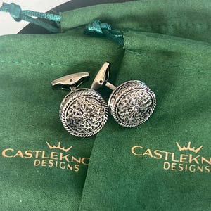 Op de afbeelding: Een paar zilverkleurige manchetknopen met een sierlijk, cirkelvormig ontwerp, rustend op een groene fluwelen zakje. De manchetknopen hebben een gedetailleerd, wervelend patroon. Op het zakje staat de tekst "CASTLEKNO DESIGNS" in goud.
