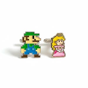 May include: A pair of silver-toned metal cufflinks featuring pixelated enamel designs. One cufflink depicts a character with a green hat and blue overalls, while the other cufflink displays a character with yellow hair and a pink dress.