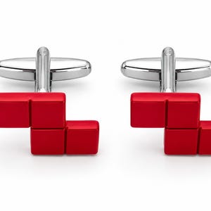 May include: A pair of silver-toned metal cufflinks featuring a bright red, blocky, L-shaped design. Each cufflink has a polished metallic toggle backing attached to a vibrant red, three-dimensional geometric shape composed of three connected squares.