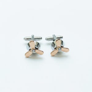 May include: A pair of cufflinks featuring a three-blade propeller design in a rose gold tone. Each propeller centre is accented with a small, clear, round-cut gemstone. The base of each cufflink is made of polished silver-tone metal.